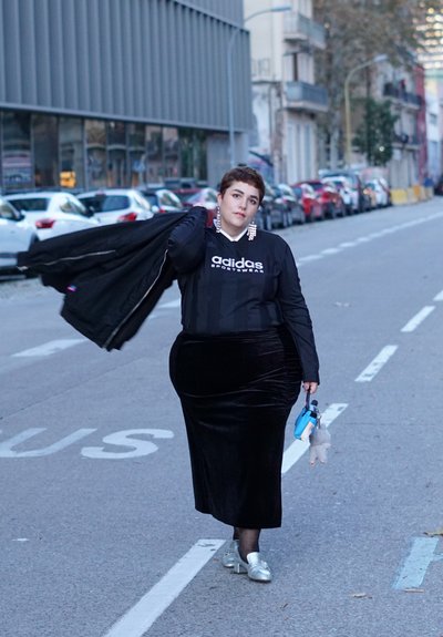 Falda de terciopelo negro, sudadera negra de Adidas con logo blanco, sosteniendo un bolso azul claro con un juguete de peluche. Zapatos y aretes plateados. Ambiente urbano.
