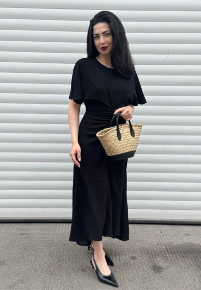 Vestido negro fluido con mangas cortas y cintura fruncida; combinado con tacones negros puntiagudos y un bolso de mano de mimbre con asas negras.
