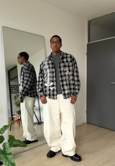 Hombre con gafas, camisa de cuadros en blanco y negro con estampado blanco, camiseta gris, pantalones anchos color crema y zapatos negros, de pie junto a un gran espejo de pie.