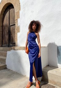 Navy satijnen jurk met ruches en een slit tot boven de dij, gecombineerd met een oranje clutch en bijpassende sleehakken op een stenen achtergrond.
