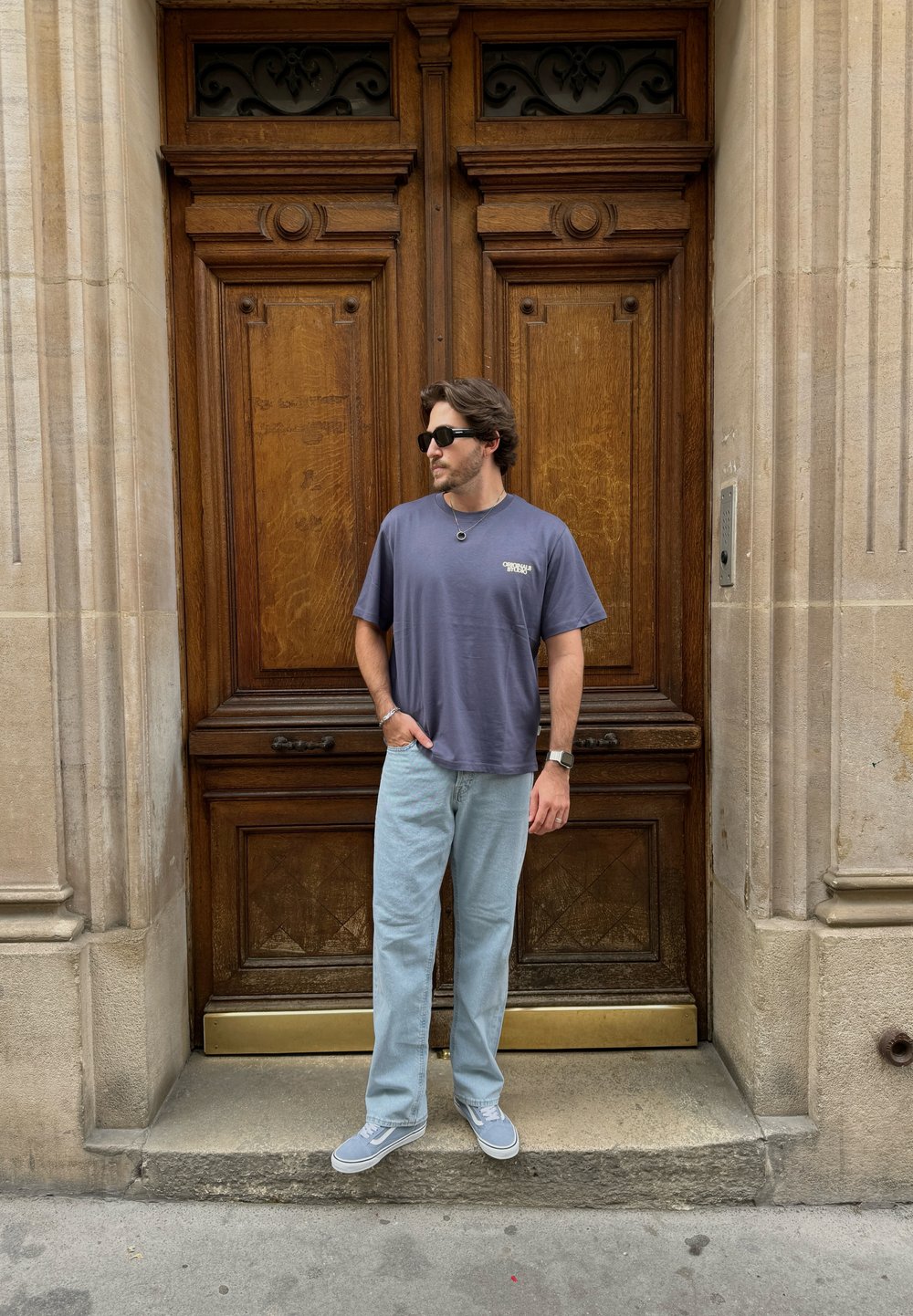 Γκρι μπλουζάκι, ανοιχτό μπλε τζιν, γκρι αθλητικά παπούτσια. Στέκεται μπροστά σε μια ξύλινη πόρτα με διακοσμητική επιφάνεια. Γυαλιά ηλίου και απλά κοσμήματα.