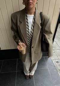 Sacou maro cu carouri mari, cămașă albă, cravată în dungi, pantaloni bej. Ținând o cafea rece, geanta maro împletită, mănuși maro.
