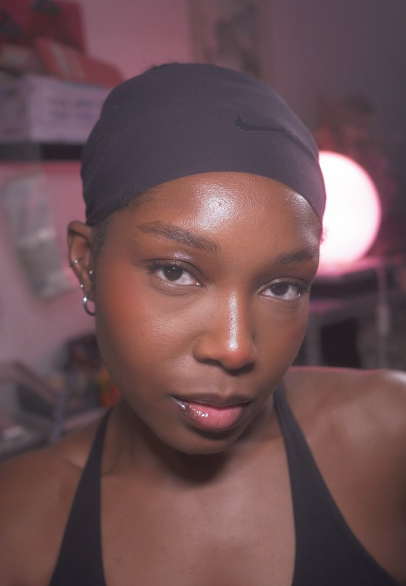 Gros plan d'une personne à la peau lisse portant un foulard noir et un débardeur, éclairée doucement par une lumière rose-violette en arrière-plan.