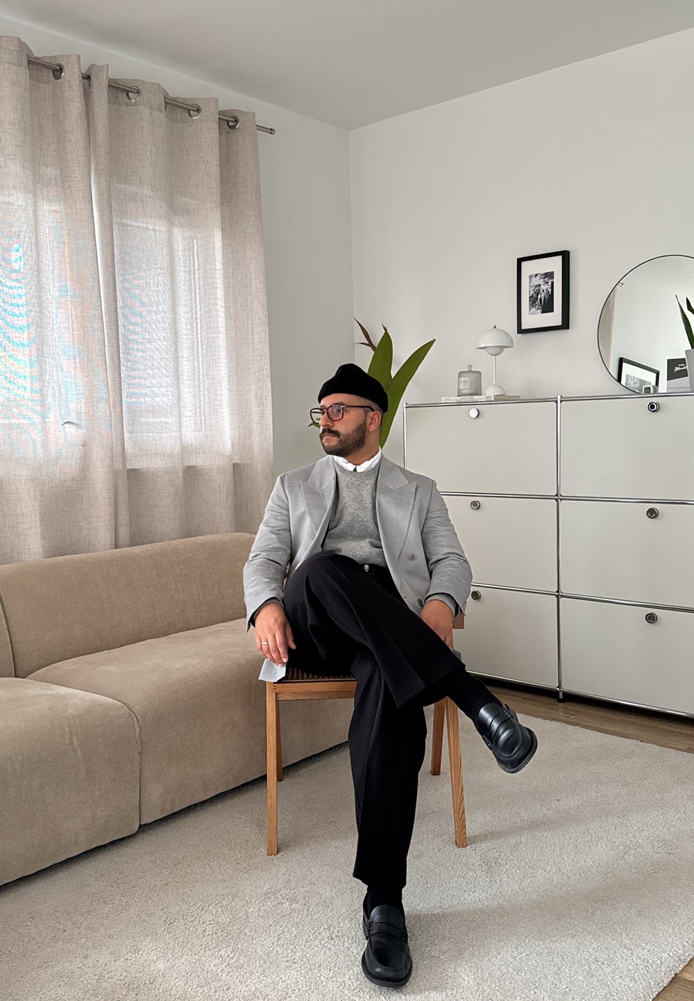 Grau-blazer über einem grauen Pullover, schwarze Hosen und Schuhe. Sitzend auf einem Holzstuhl neben einem beigen Sofa, mit hellen Vorhängen und Pflanzen.