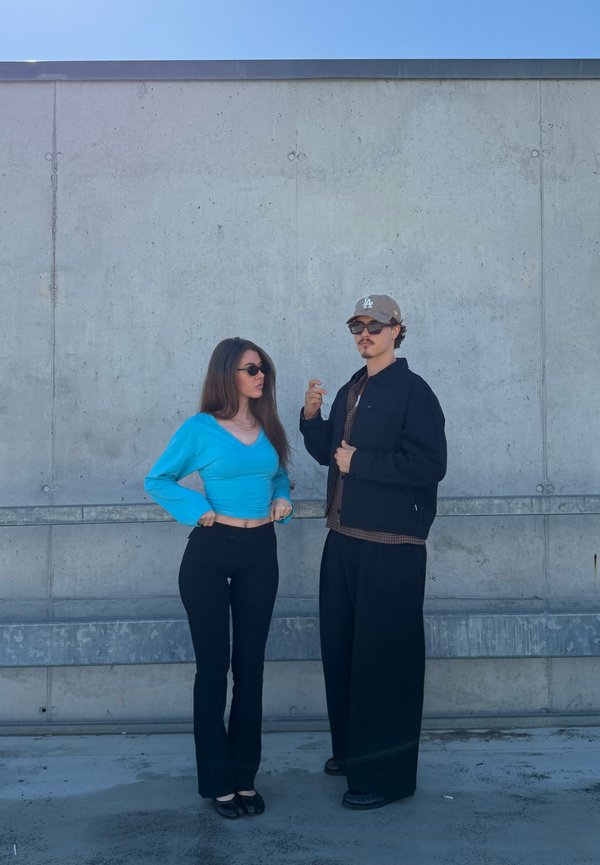 Giovane uomo e donna con occhiali da sole sono appoggiati a un muro di cemento, la donna con una canotta blu e l'uomo con abiti casual scuri e un cappello.