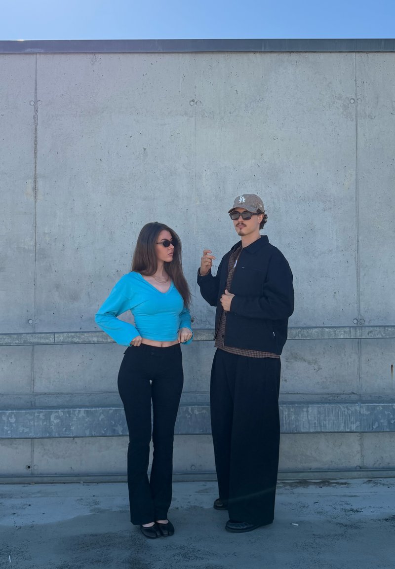 Jonge man en vrouw met een zonnebril staan tegen een betonnen muur, de vrouw in een blauw topje en de man in donkere casual kleding en een pet.