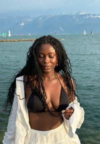 Svart triangulär bikiniöverdel, genomskinlig vit cover-up draperad över ena axeln, lång lockig hår, sjö bakgrund med berg och segelbåtar.