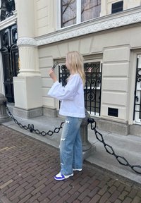 Camisa blanca de gran tamaño con mangas largas, jeans desgastados de color azul claro con detalles de lazo blanco, y zapatillas con acentos morados en un camino de adoquines.