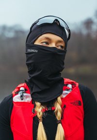 Personne aux cheveux blonds tressés portant un cagoule noire, des lunettes de soleil et un gilet d'hydratation rouge en plein air dans un environnement brumeux.