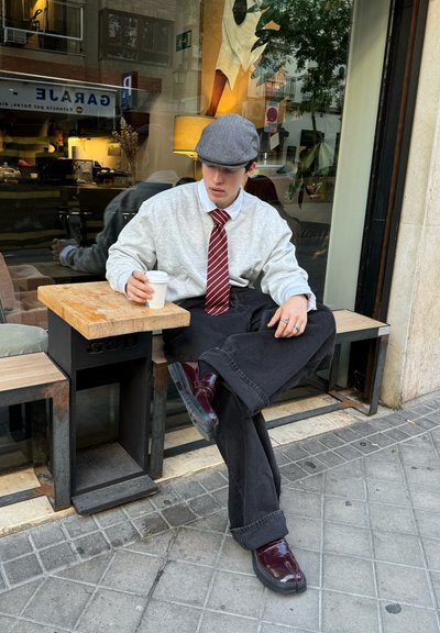 Sudadera gris, camisa blanca, corbata a rayas rojas, pantalones de pierna ancha negros y zapatos burdeos brillantes, sentado en una mesa de madera al aire libre.