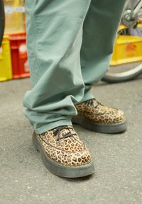 Leopardimustriga platvormijalatsid mustade kummitaldadega, nööpide ja õmblusdetailidega, koos lõtvade roheliste pükstega.