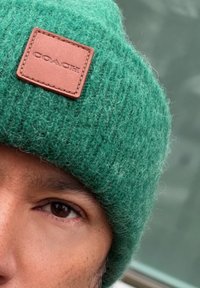 Gorra de lana verde de punto con un acabado texturizado, que presenta un parche rectangular de cuero marrón con la palabra "COACH" en relieve.