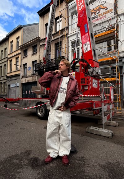 Joven con chaqueta roja, pantalones blancos y zapatos rojos está de pie en la calle cerca de una grúa de construcción roja y andamios en la fachada de un edificio.
