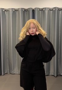 Haut noir en maille avec de larges manches, assorti à un short noir. Cheveux blonds longs et ondulés, sur fond de rideaux gris et sol carrelé.