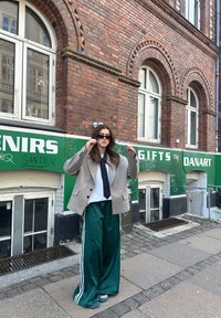 Νέα γυναίκα που φορά γυαλιά ηλίου, υπερμεγέθη σακάκι, λευκό πουκάμισο, μαύρη γραβάτα και φαρδιά πράσινα παντελόνια στέκεται στο πεζοδρόμιο της πόλης μπροστά από κτίριο με τούβλα και πράσινη πινακίδα.