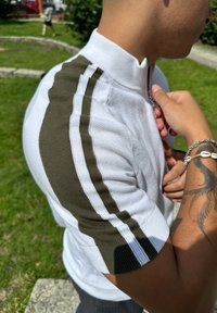 Polo shirt bianca con colletto zip, presenta dettagli a righe verde oliva e bianche sulle maniche. Tessuto texturizzato, design aderente.