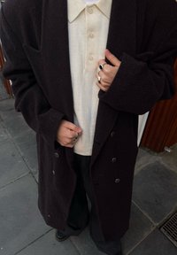 Manteau texturé sombre avec boutons argentés, porté sur un polo crème. Les mains du mannequin sont ornées de plusieurs bagues en argent. Pantalon noir visible.