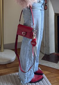 Red leather crossbody bag with a front flap and silver hardware, featuring keychains including a heart tag, worn with light blue jeans.