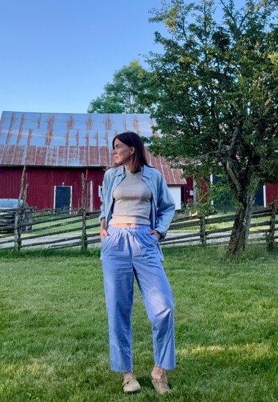 Camisa azul claro, tipo polo, sobre una camiseta gris, combinada con pantalones sueltos a rayas en azul y blanco. El entorno incluye un área de césped y un granero.