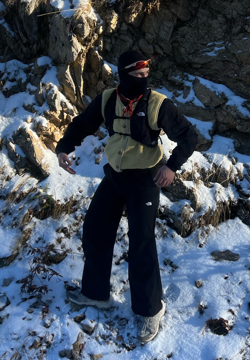 Personne portant une cagoule noire, un gilet beige et des vêtements noirs, debout sur un terrain rocheux enneigé, regardant sur le côté avec les bras légèrement pliés.