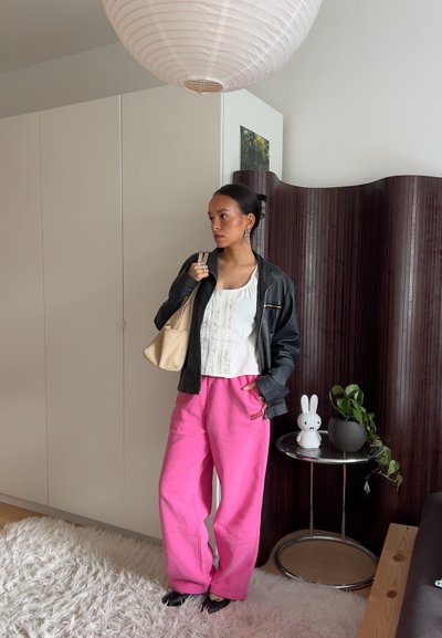 Mujer de pie en el interior con chaqueta negra, blusa blanca, pantalones rosa brillante, sosteniendo una bolsa beige, cerca de una pequeña mesa con lámpara de conejo y planta.