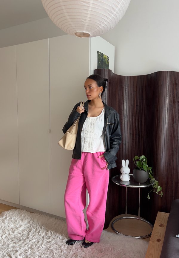 Femme debout à l'intérieur portant une veste noire, un chemisier blanc, un pantalon rose vif, tenant un sac beige, près d'une petite table avec une lampe lapin et une plante.