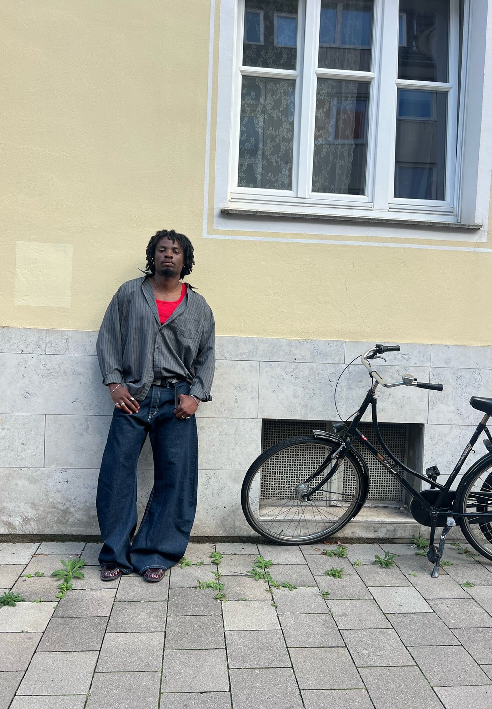 Czarny rower obok osoby ubraną w szarą koszulkę w paski na czerwonym tle i luźne niebieskie dżinsy, stojącej na chodniku obok roślin.