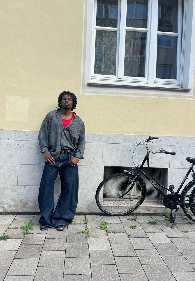 Schwarzes Fahrrad neben einer Person, die ein gestreiftes graues Shirt über einem roten Oberteil und weite blaue Jeans trägt, auf einem Bürgersteig mit Pflanzen stehend.