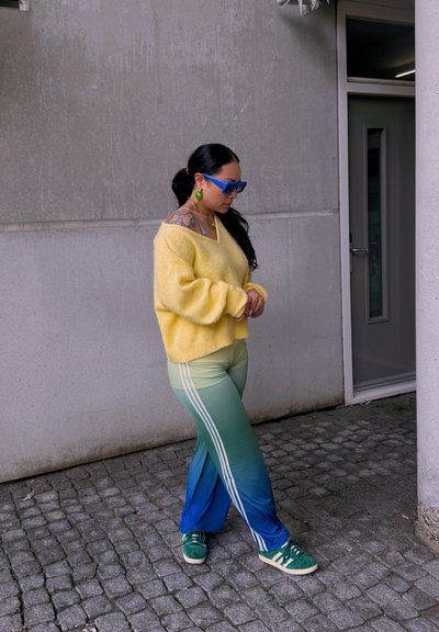 Suéter amarillo esponjoso, pantalones deportivos verdes con degradado y rayas blancas, zapatillas verdes, gafas de sol azules y grandes pendientes verdes.