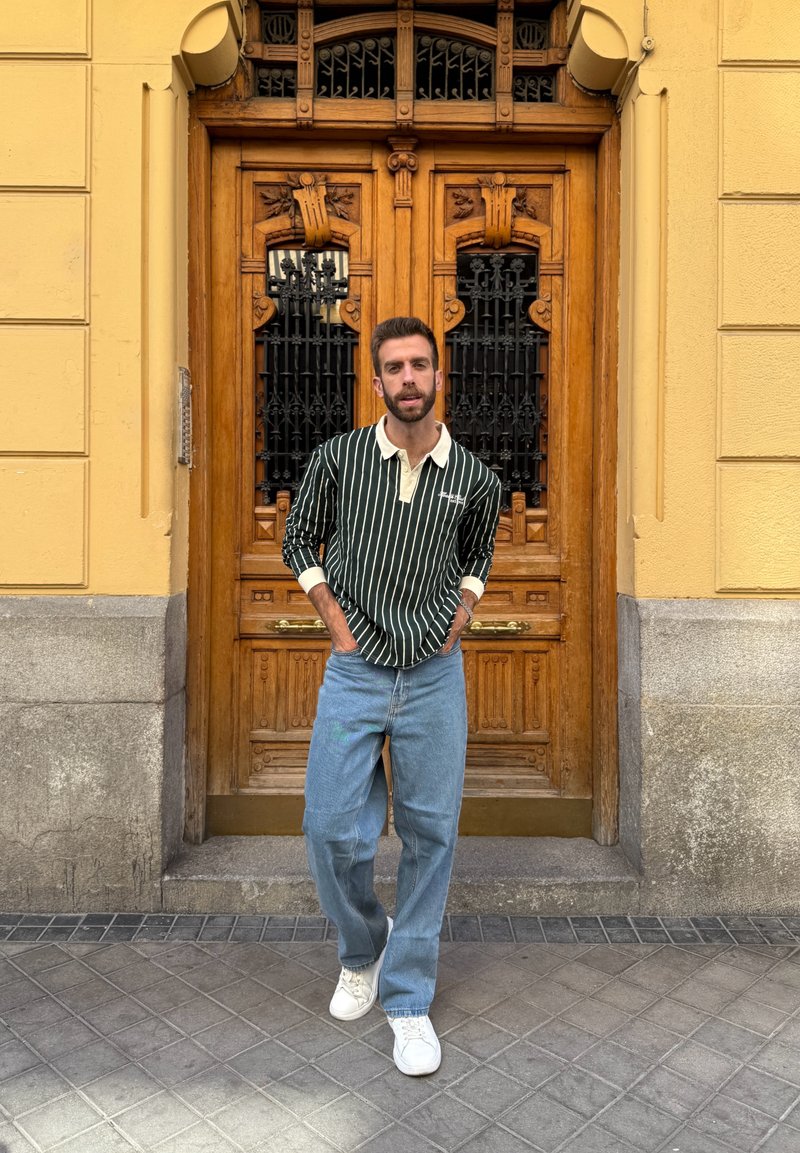 Grün-weiß gestreiftes Langarmhemd mit Kragen, kombiniert mit hellblauen Jeans und weißen Sneakers. Holztür im Hintergrund.
