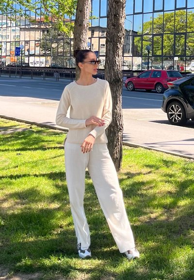 Mujer con suéter crema y pantalones con zapatillas negras de pie sobre el césped junto a dos árboles, calle de la ciudad y coches aparcados al fondo.