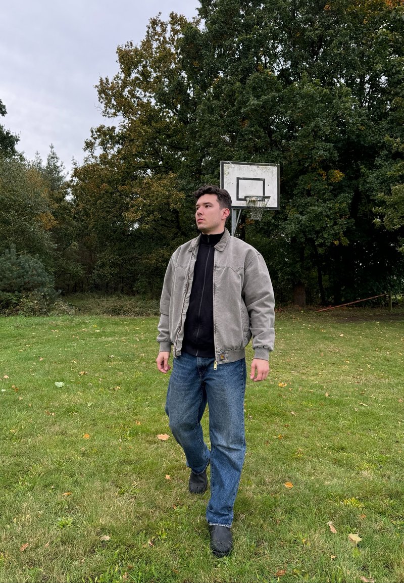 Γκρι bomber jacket πάνω από μια μαύρη hoodie, συνδυασμένο με μπλε τζιν παντελόνι. Στο φόντο υπάρχει μια εξωτερική μπασκέτα ανάμεσα σε δέντρα. Γκαζόν από κάτω.