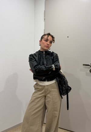 Persona joven con cabello rizado, chaqueta de cuero negra y pantalones beige, de pie con los brazos cruzados cerca de una puerta cerrada en un interior.