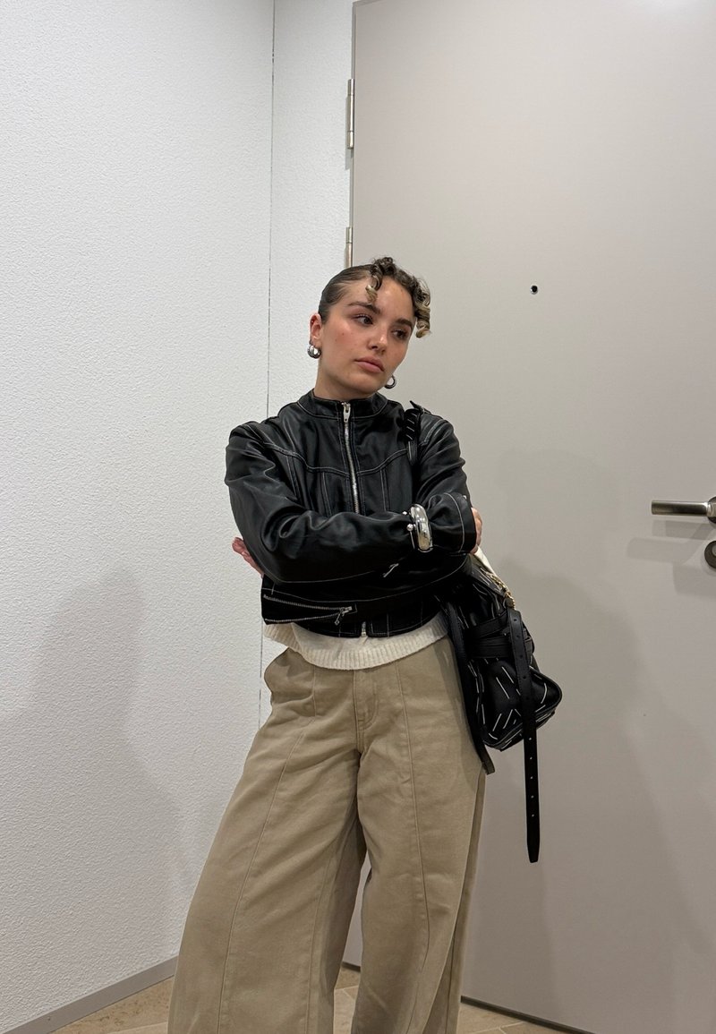 Ung person med krøllete hår iført svart skinnjakke og beige bukser står med armene krysset ved en lukket dør innendørs.