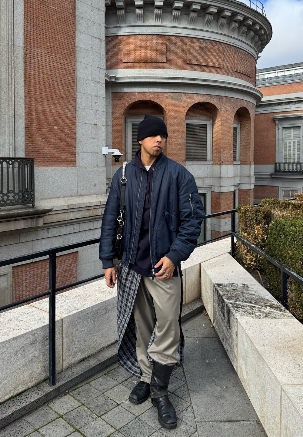 Ναυτική bomber τζάκετ, μαύρο καπέλο beanie, μαύρη τσάντα crossbody, γκρι σατέν παντελόνι με καρό λεπτομέρειες, μαύρες μποτες με ύψος μέχρι το γόνατο, με φόντο το τούβλινο αρχιτεκτονικό στυλ.