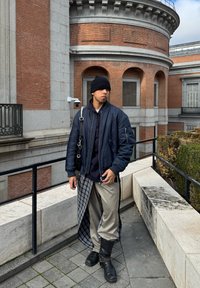 Ναυτική bomber τζάκετ, μαύρο καπέλο beanie, μαύρη τσάντα crossbody, γκρι σατέν παντελόνι με καρό λεπτομέρειες, μαύρες μποτες με ύψος μέχρι το γόνατο, με φόντο το τούβλινο αρχιτεκτονικό στυλ.