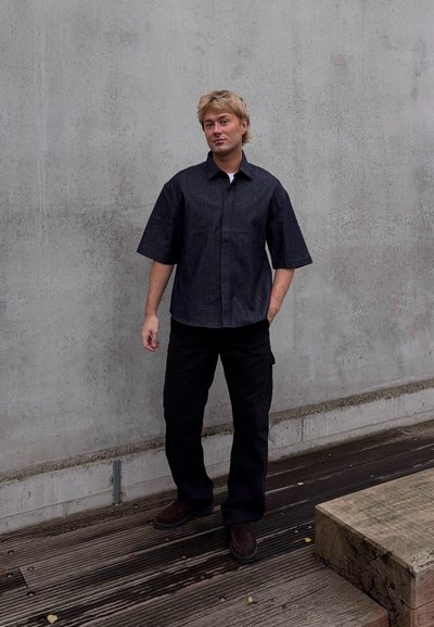 Camisa de denim oscuro de manga corta con cuello abotonado, combinada con pantalones negros y calzado oscuro, de pie sobre una tarima de madera contra una pared gris.