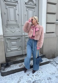 Femme portant une veste en fausse fourrure rose, un chemisier à nœud, un jean à jambes larges et des bottes pointues, tenant une pochette matelassée marine sur des marches enneigées près d'une porte ornée.