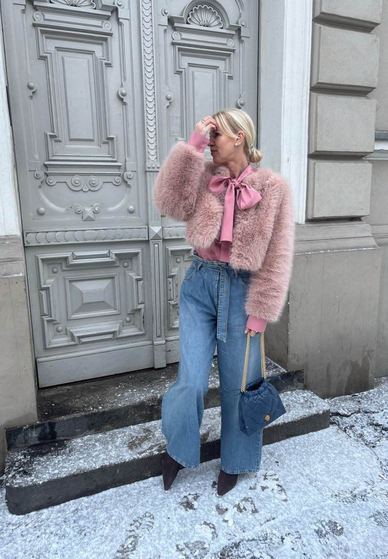 Frau in rosa Kunstpelzjacke, gebundener Bluse, weit geschnittenen Jeans und spitzen Stiefeln, die eine marineblaue gesteppte Tasche auf verschneiten Stufen neben einer verzierten Tür hält.