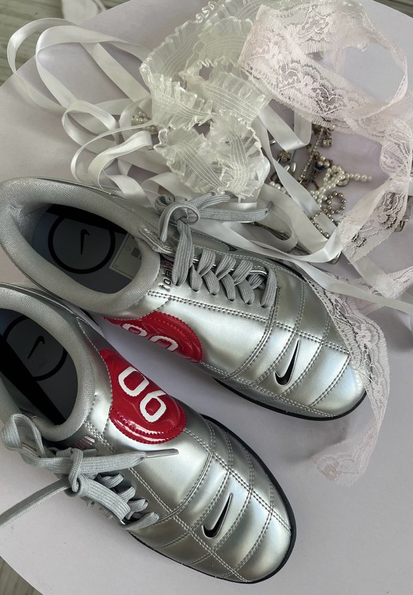 Zapatillas Nike plateadas con detalles rojos junto a cintas de encaje blancas y joyas de perlas sobre una superficie clara.