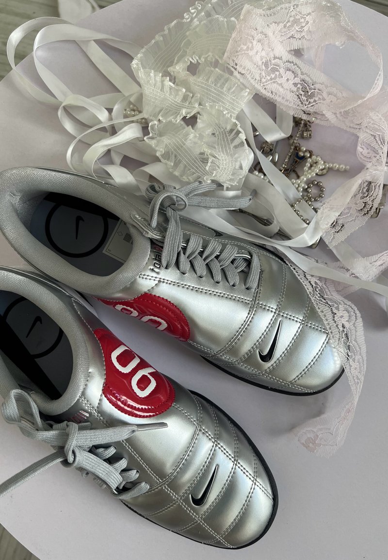 Baskets Nike argentées avec des accents rouges à côté de rubans en dentelle blancs et de bijoux en perles sur une surface claire.