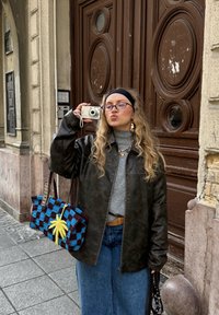 Pruun nahktagi, helehall rullkaelusega kampsun, teksaseelik. Sinise ja pruuni ruuduline kott kollase aktsendiga. Tellissein taustal.