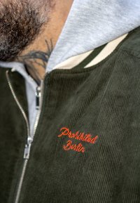 Veste en velours côtelé verte avec l'inscription brodée "Prohibited Berlin" en orange. Dotée d'une fermeture éclair et d'un sweat à capuche gris clair en dessous.