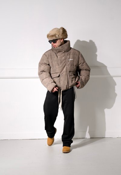 Chaqueta acolchada marrón con un acabado texturizado, cuello alto y dos bolsillos frontales. Combinada con pantalones negros y zapatos deslizables color beige, además de un gorro de pelo.