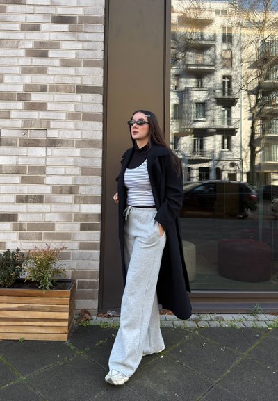 Mujer con abrigo largo negro, camiseta de rayas, pantalones anchos grises, zapatillas deportivas y gafas de sol, de pie en una acera urbana cerca de una pared de ladrillos y una ventana de cristal.