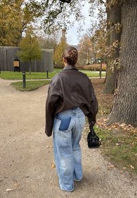 Brun läderoversized jacka, ljusblå baggy jeans med en lappficka och en liten svart handväska. Utomhusmiljö med träd i bakgrunden.