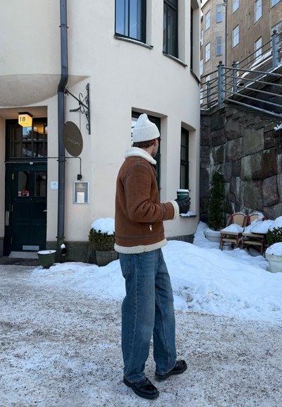 Hombre con chaqueta marrón de borrego y gorro blanco sosteniendo una taza de café de pie en un patio nevado cerca de un edificio beige y un muro de piedra.