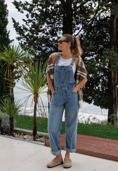 Overoles de mezclilla en azul claro, combinados con una camiseta de tirantes blanca y un cárdigan de punto marrón, complementados con zapatos tipo slip-on en beige claro.