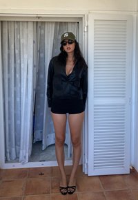 Čierna skrátená kožená bunda s dvoma prednými vreckami, čierne šortky, zelená čiapka s bielym logom, okrúhle slnečné okuliare a čierne sandále na viazanie.