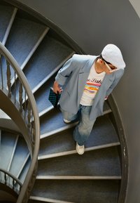 Una persona sale su una scala a chiocciola indossando un blazer azzurro chiaro, una maglietta grafica, jeans larghi e sneakers bianche, mentre tiene in mano una pochette a fantasia.